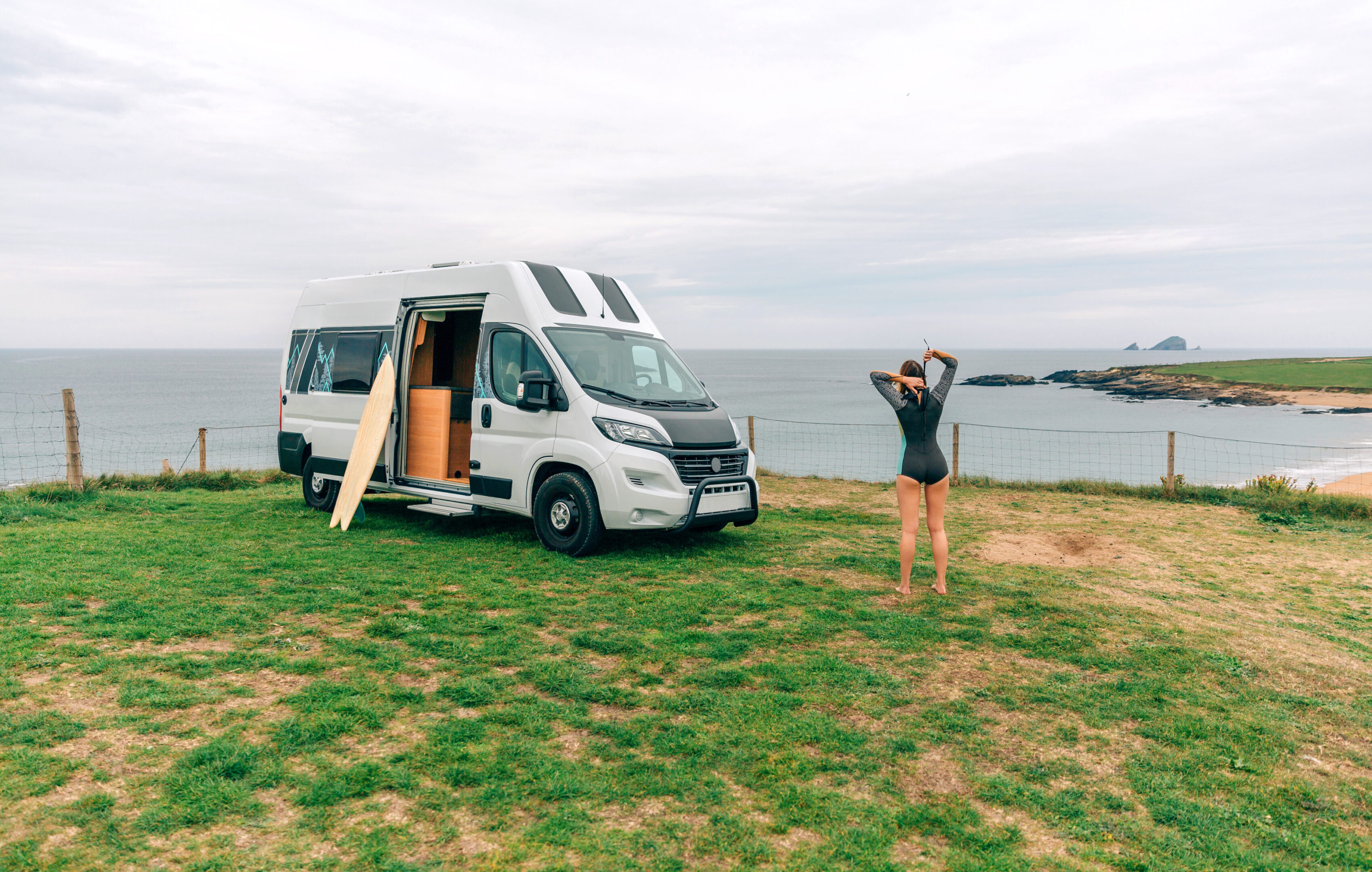Unrecognizable young woman closing the zipper of her wetsuit for surfing next to her camper van